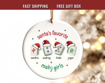 May include: A round white ceramic ornament featuring four mahjong tiles, each topped with a red and white Santa hat. The text reads Santa's favorite mahj girls, with the names sandra, audrey, linda, and joyce printed below each tile.