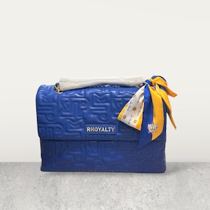 Puede incluir: Un bolso azul real con un patrón acolchado y la palabra "RHOYALTY" en la parte delantera. Una bufanda decorativa en blanco, amarillo y azul está atada al asa. El bolso tiene una correa de cadena dorada.