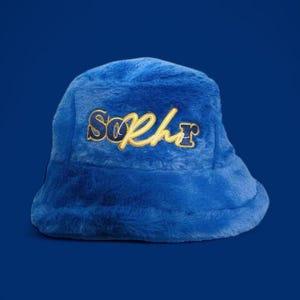 May include: A plush, blue bucket hat with the word "SoPhr" embroidered in gold thread. The hat has a soft, fuzzy texture and a rounded crown. The background is a solid, darker blue.