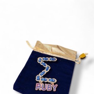 May include: A gold-coloured metal brooch in a zigzag design with blue detailing, on a navy blue velvet pouch. The brooch has the word "RUBY" in red and blue. The pouch has a gold drawstring.