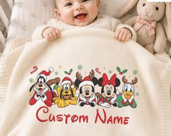 May include: A cream-colored knit baby blanket featuring an embroidered graphic of Disney characters including Goofy, Pluto, Mickey Mouse, Minnie Mouse, and Donald Duck in holiday attire, with the red embroidered text Custom Name below them.