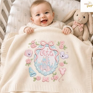 May include: A cream-colored knit baby blanket featuring an embroidered design of a castle inside a blue oval frame, the name Olivia, a pink bow, roses, a wizard hat, a camera, and mouse-shaped icons. A plush tan rabbit toy sits nearby.