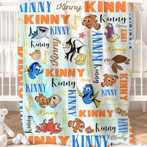 May include: A blue and orange baby blanket with a Finding Nemo theme. The blanket features the characters Dory, Nemo, Marlin, and other sea creatures. The name "Kinny" is repeated throughout the blanket.