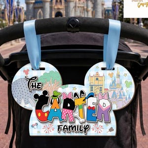 May include: A stroller sign shaped like mouse ears with a light blue checkered background. It features the text THE CARTER FAMILY, illustrations of theme park landmarks, and colorful character-themed letters. Two light blue ribbons attach it.