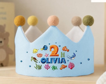 May include: A light blue fabric crown featuring a large orange number 2 and the name OLIVIA in dark blue letters. The design includes embroidered sea creatures like fish, a turtle, and a stingray, with colorful coral and five felt pom-poms on top.