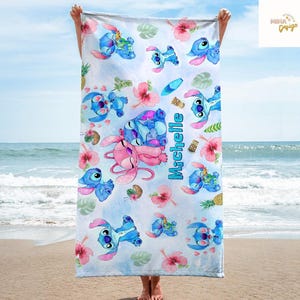 May include: A beach towel featuring a repeating pattern of blue and pink cartoon characters, tropical flowers, and pineapples on a light blue background. The name "Michelle" is printed vertically in turquoise. The towel is held up against a beach backdrop.