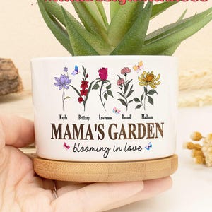 May include: White ceramic planter with a floral design and the text "Mama's Garden blooming in love". The planter has a bamboo base and features five flowers with names written below each flower.