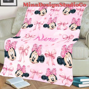 Puede incluir: Manta de forro polar rosa con un patr&oacute;n repetido de cabezas de Minnie Mouse y lazos rosas. La manta tiene la palabra "Name" escrita en cursiva blanca.