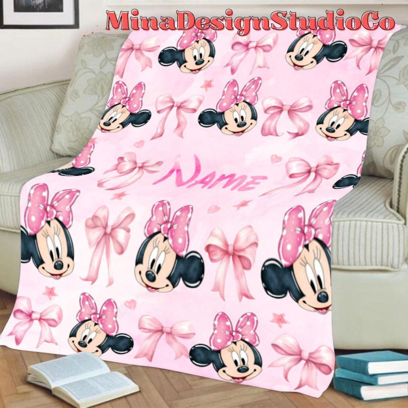 Puede incluir: Manta de forro polar rosa con un patr&oacute;n repetido de cabezas de Minnie Mouse y lazos rosas. La manta tiene la palabra "Name" escrita en cursiva blanca.