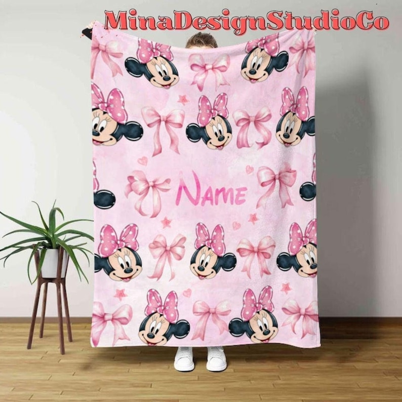 Puede incluir: Manta de forro polar rosa con un patr&oacute;n repetido de cabezas de Minnie Mouse y lazos rosas. La manta tiene la palabra "NAME" impresa en el centro.