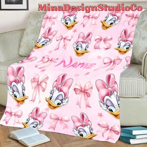 Puede incluir: Manta de forro polar rosa con un patr&oacute;n repetido de un pato de dibujos animados con un lazo rosa y lazos rosas. El texto "Name" est&aacute; impreso en la manta.