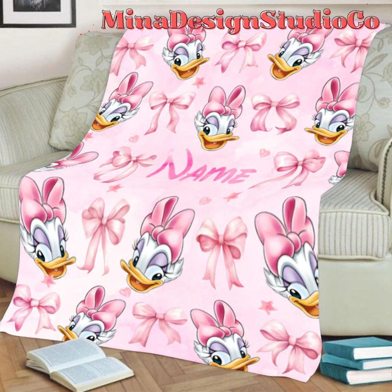 Puede incluir: Manta de forro polar rosa con un patr&oacute;n repetido de un pato de dibujos animados con un lazo rosa y lazos rosas. El texto "Name" est&aacute; impreso en la manta.