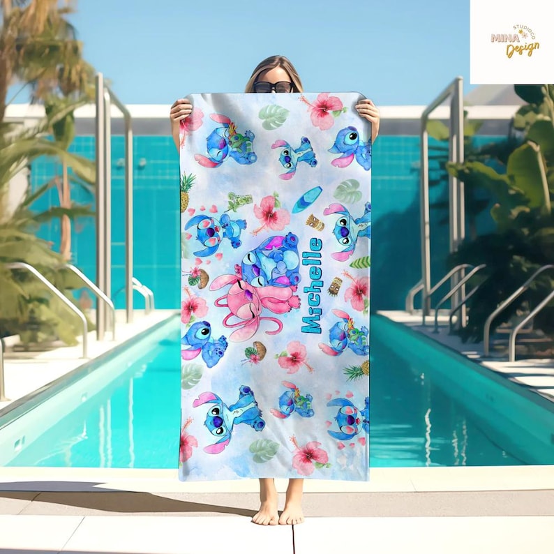 May include: A light blue and white beach towel featuring cartoon characters, pink flowers, and tropical elements. The towel has the name "Michelle" printed in blue. The design includes images of a blue alien-like creature and a pink alien-like creature.