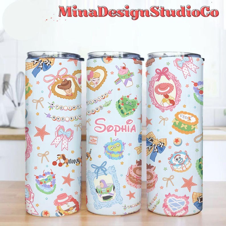 K&ouml;nnte beinhalten: Drei Edelstahlbecher mit hellblauem Hintergrund und verschiedenen bunten Comicfiguren und Designs. Die Becher zeigen eine rosa-wei&szlig;e Schleife, einen gr&uuml;nen Au&szlig;erirdischen, eine Toy-Story-Figur und einen Kuchen. Der Text "Sophia" ist auf einem der Becher aufgedruckt.