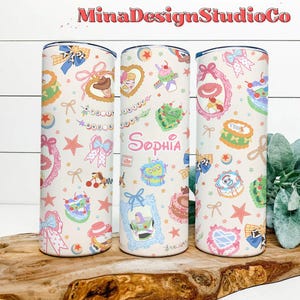 K&ouml;nnte beinhalten: Drei Edelstahlbecher mit wei&szlig;em Hintergrund und einem bunten Muster aus Comicfiguren, Schleifen, Sternen und dem Namen "Sophia".