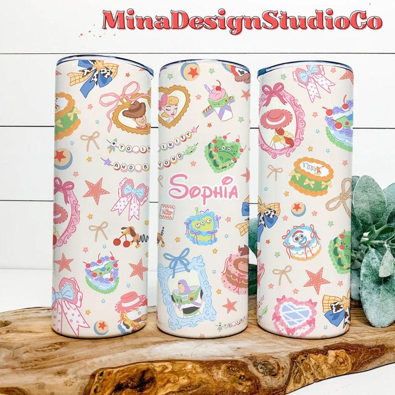 K&ouml;nnte beinhalten: Drei Edelstahlbecher mit wei&szlig;em Hintergrund und einem bunten Muster aus Comicfiguren, Schleifen, Sternen und dem Namen "Sophia".