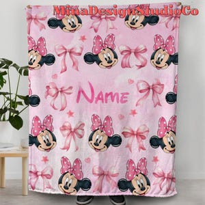 Puede incluir: Manta de forro polar rosa con un patr&oacute;n repetido de cabezas de Minnie Mouse y lazos rosas. La manta tiene la palabra "Nombre" impresa en el centro.