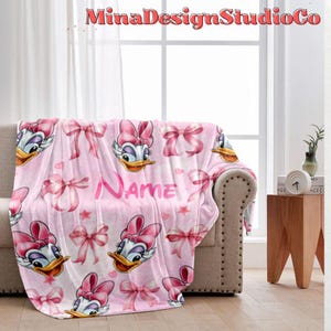 Puede incluir: Manta de forro polar rosa con un patr&oacute;n repetido de un pato de dibujos animados con un lazo rosa, lazos rosas y estrellas. La manta tiene la palabra "Nombre" impresa en ella.