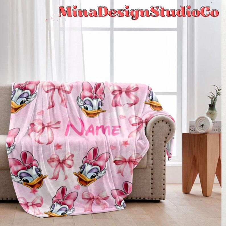 Puede incluir: Manta de forro polar rosa con un patr&oacute;n repetido de un pato de dibujos animados con un lazo rosa, lazos rosas y estrellas. La manta tiene la palabra "Nombre" impresa en ella.