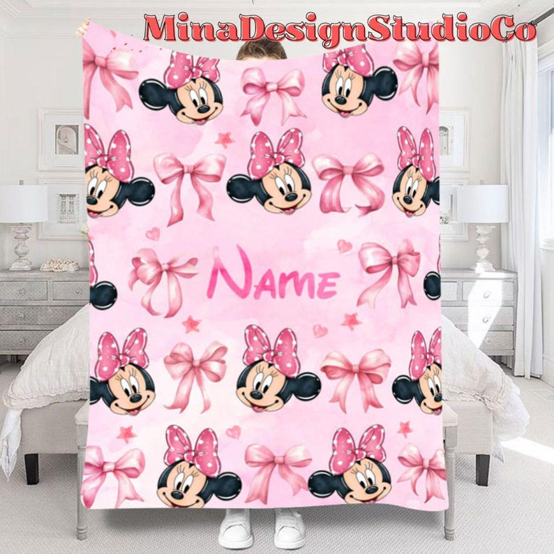 Puede incluir: Manta de forro polar rosa con un patr&oacute;n repetido de cabezas de Minnie Mouse, lazos rosas y estrellas. La manta tiene la palabra "Name" en el centro.