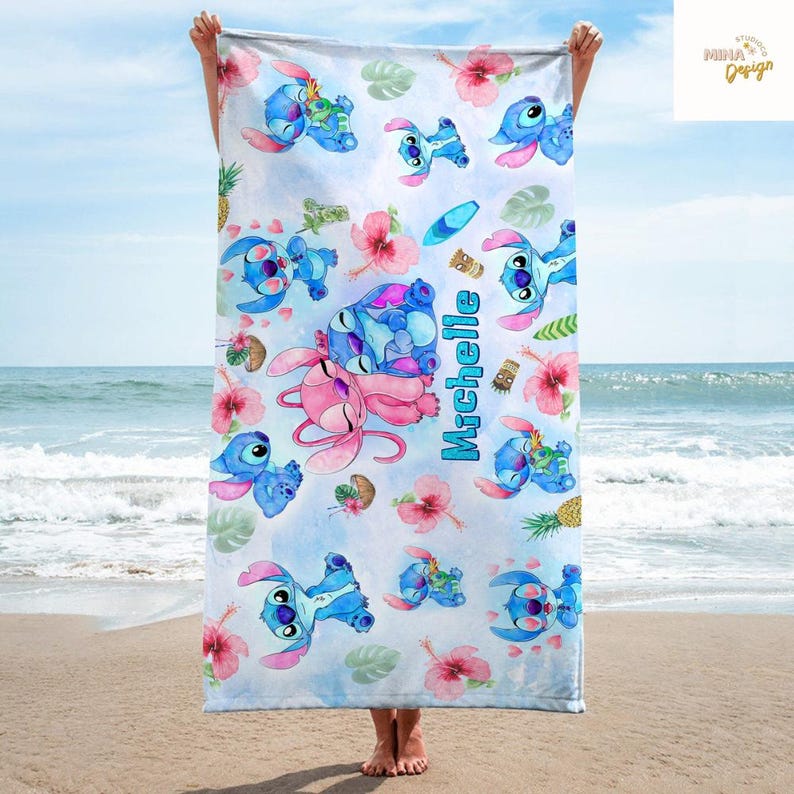 May include: A beach towel featuring a repeating pattern of blue and pink cartoon characters, tropical flowers, and pineapples on a light blue background. The name "Michelle" is printed vertically in turquoise. The towel is held up against a beach backdrop.