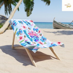 May include: A beach towel featuring a blue and white pattern with cartoon characters, flowers, and the name "Michelle". The towel is draped over a wooden beach chair on a sandy beach with a palm tree and a boat in the background.
