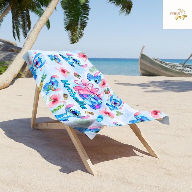 May include: A beach towel featuring a blue and white pattern with cartoon characters, flowers, and the name "Michelle". The towel is draped over a wooden beach chair on a sandy beach with a palm tree and a boat in the background.