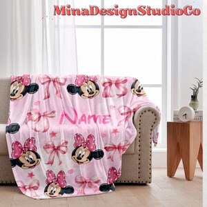 Puede incluir: Manta de forro polar rosa con un patr&oacute;n repetido de cabezas de Minnie Mouse, lazos rosas y estrellas. La manta tiene la palabra "Name" impresa en letras rosas.