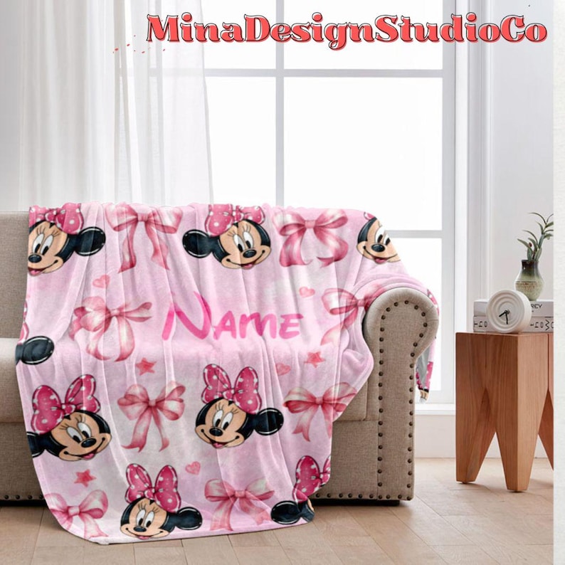 Puede incluir: Manta de forro polar rosa con un patr&oacute;n repetido de cabezas de Minnie Mouse, lazos rosas y estrellas. La manta tiene la palabra "Name" impresa en letras rosas.
