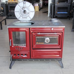Könnte beinhalten: Ein freistehender roter Holzofen mit metallischer Oberfläche, einer verglasten Feuerbox auf der linken Seite und einem Backofen rechts mit integriertem Thermometer. Der Ofen hat einen runden Deckel, der über der Kochfläche angehoben ist.