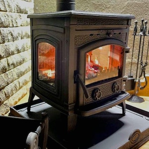 May include: A black cast iron wood-burning stove with a glass door and a fire burning inside. The stove is sitting on a black metal base.