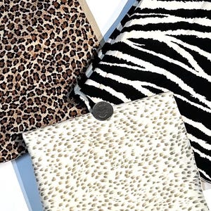 May include: An assortment of fabric swatches featuring animal prints. The swatches include a leopard print in brown and black, a zebra print in black and white, and a speckled print in beige and brown. A quarter coin is included for scale.
