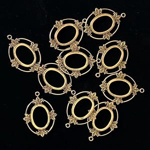 May include: A collection of gold-colored oval pendant frames with leaf accents. Each frame has an open center and small loops for attaching to other jewelry components. The frames are scattered on a black background.