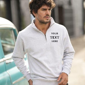 Puede incluir: Una sudadera gris con cremallera de media cremallera con un diseño bordado en negro que dice "YOUR TEXT HERE".