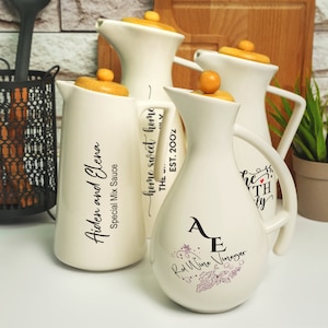 Op de afbeelding: Vier witte keramische flessen met houten deksels en zwarte letters. De flessen zijn gelabeld met "Aiden en Elena Special Mix Sauce", "home sweet home THE EST. 2002" en "Rode wijnazijn".