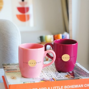 May include: Two ceramic mugs with a pink and burgundy color scheme. Both mugs feature a gold circle with the text "JAMIE & ANNIE STUDIO" in black.