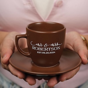 以下が含まれることがあります： 白い文字で「mr & mrs ROBERTSON EST. 05.25.2024」と書かれた、茶色のセラミック製のコーヒーカップとソーサーのセットです。