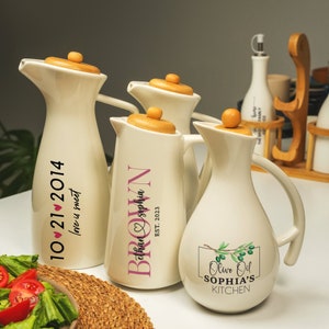 May include: Several white ceramic oil and vinegar dispensers with wooden lids are displayed. Some have custom text in black and pink, including dates and names. One dispenser features an olive branch design and the text "Olive Oil SOPHIA'S KITCHEN."