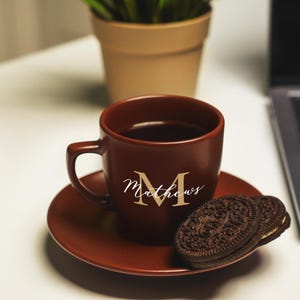 Puede incluir: Una taza de café de cerámica marrón con el nombre "Matthews" escrito en letras doradas. La taza está llena de café y está sobre un platillo marrón con dos galletas de chocolate.