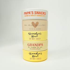 Puede incluir: Una pila de cuencos de cerámica personalizados en varios colores. El cuenco superior es blanco con texto naranja que dice "PAPA'S SNACKS". Otros cuencos son de color beige y amarillo, con texto como "Grandpa's Bowl" y nombres.