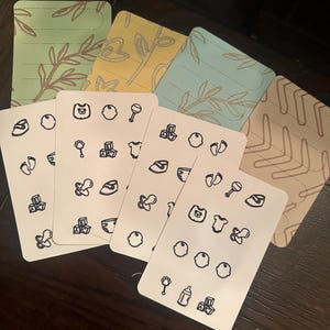 May include: A collection of baby-themed cards with various illustrations. The cards feature black line drawings of baby items like bottles, pacifiers, and toys on a white background. The backs of the cards have pastel floral designs in green, yellow, and blue.