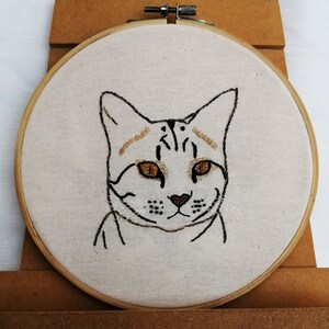 May include: Hand-stitched embroidery art featuring a cat's face. The cat has brown eyes, a brown nose, and brown and black markings. The embroidery is set in a wooden hoop on a cream-colored fabric.