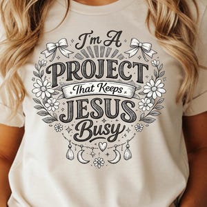 May include: A beige t-shirt with the text "I'm A PROJECT That Keeps JESUS Busy" in a floral wreath design. The shirt has short sleeves and is paired with blue jeans. The word "NATURAL" is printed in a black rectangle at the bottom.