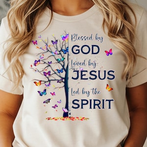 Può includere: T-shirt beige con un disegno ad albero e la scritta "Blessed by GOD, Loved by JESUS, Led by the SPIRIT." L'albero presenta farfalle colorate. La parola "NATURAL" è stampata in basso nell'immagine.