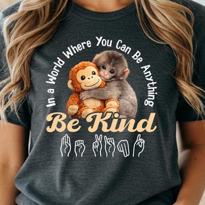 May include: Dark heather gray t-shirt with a graphic of two hugging monkeys and the words "In a World Where You Can Be Anything, Be Kind" in white. The shirt also features sign language symbols for "Be Kind".