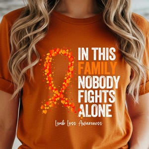 Puede incluir: Una camiseta naranja con el texto "IN THIS FAMILY NOBODY FIGHTS ALONE" en blanco, y una cinta hecha de corazones naranjas y amarillos. Las palabras "Limb Loss Awareness" están debajo del texto. La palabra "AUTUMN" está en la parte inferior izquierda.