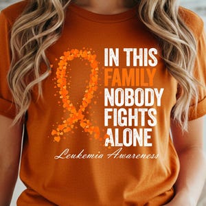 Könnte beinhalten: Ein kurzärmeliges T-Shirt in gebranntem Orange mit einer Grafik eines orangefarbenen Bewusstseinsbandes aus kleinen Herzen. Der Text IN THIS FAMILY NOBODY FIGHTS ALONE und Leukemia Awareness ist in weißer und oranger Schrift aufgedruckt.