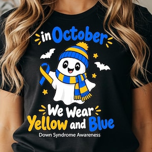 Op de afbeelding: Een zwart t-shirt met een witte geestfiguur die een blauw en geel gestreepte muts en sjaal draagt. De afbeelding bevat de tekst "in October We Wear Yellow and Blue" en "Down Syndrome Awareness" met gele sterren en vleermuizen.