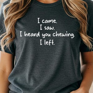 May include: A dark heather gray short-sleeve t-shirt featuring white text that reads: I came. I saw. I heard you chewing. I left. The shirt has a crew neckline and is paired with blue denim jeans that have a visible metal button and pocket rivets.