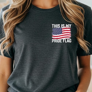 以下が含まれることがあります： 「THIS IS MY PRIDE FLAG」の文字とアメリカ国旗のデザインが施された、ダークヘザーグレーのTシャツ。クルーネックのスタイルです。